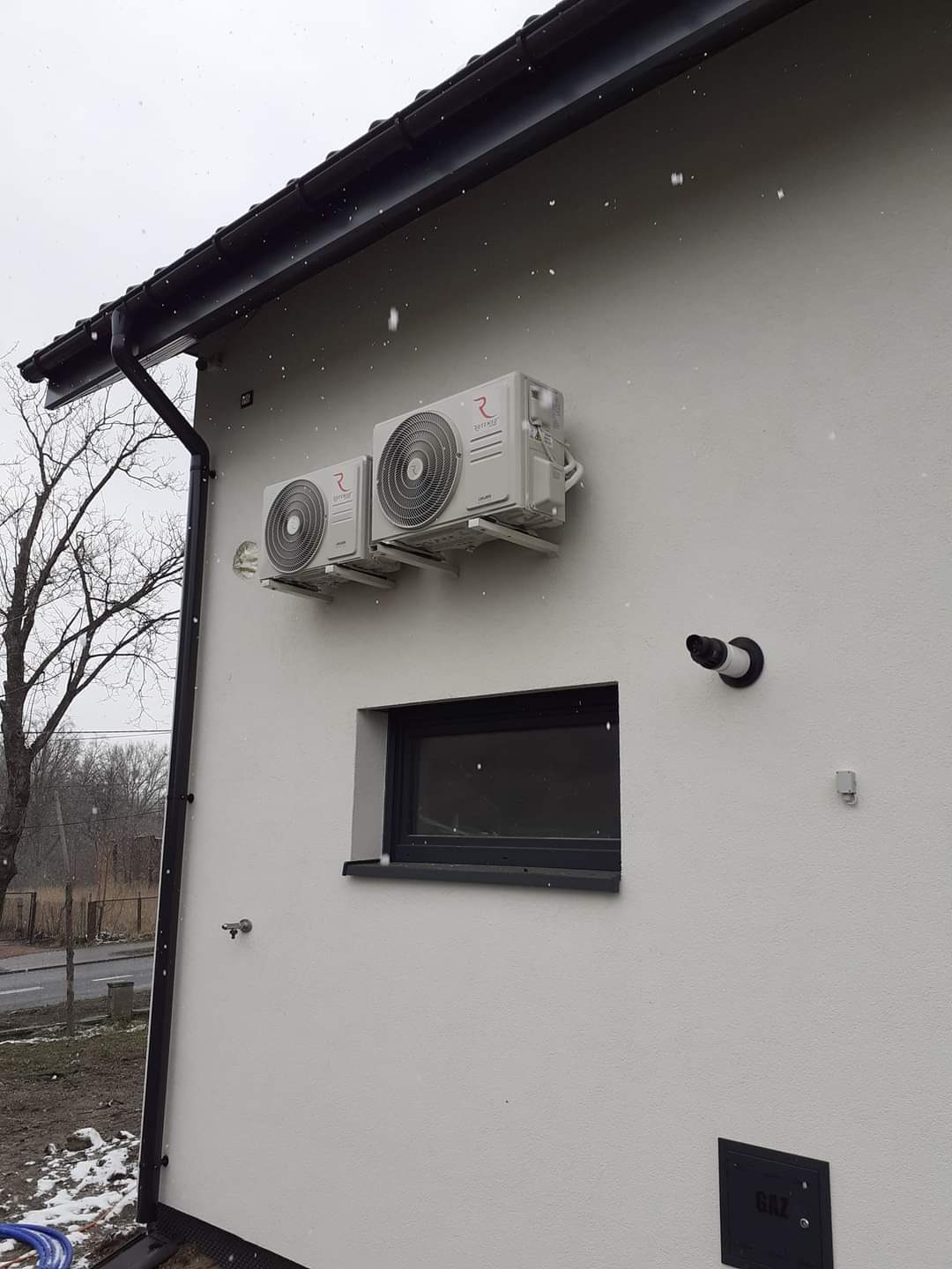 Dwa klimatyzatory zamontowane na zewnętrznej ścianie domu, widoczne rury odprowadzające skropliny, na zewnątrz budynku widać śnieg.