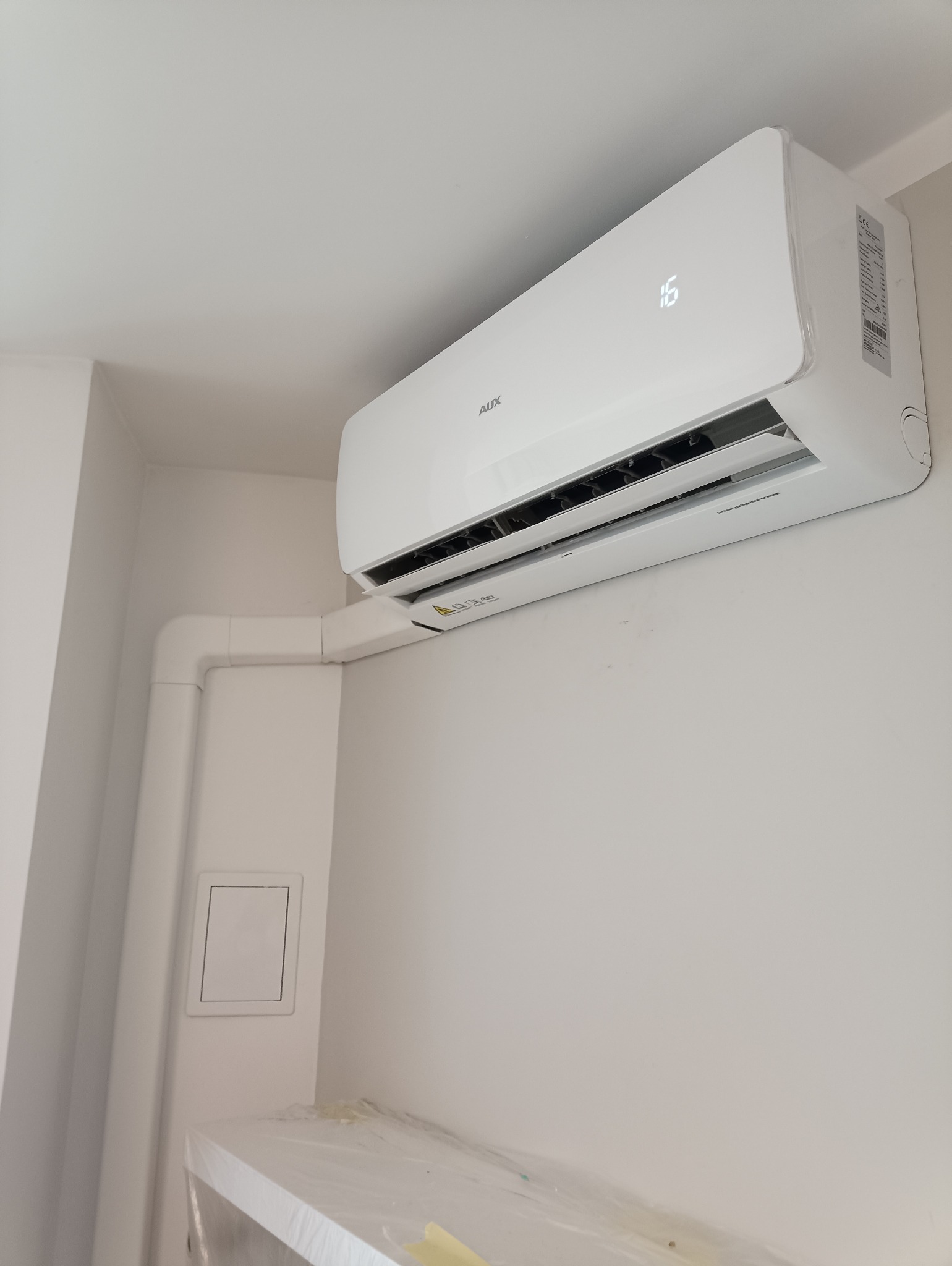 Biały klimatyzator AUX zamontowany na ścianie w pomieszczeniu, widoczne rury instalacyjne i wyświetlacz temperatury ustawiony na 16 stopni.