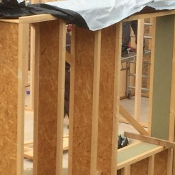 Michał Kędzierski - Konstrukcja ścian szkieletowych w trakcie montażu, widoczne drewniane ramy i płyty OSB, fragment izolacji w oknie, w tle drabina i osoba na podwyższeniu.