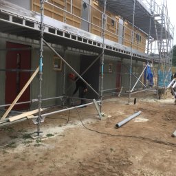 Michał Kędzierski - Budowa domu szkieletowego w trakcie: pracownik obrabia ścianę zewnętrzną spod rusztowania, widoczne drewniane elementy konstrukcyjne i narzędzia pracy.