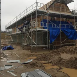 Budowa domu szkieletowego w trakcie: widoczne rusztowania, konstrukcja drewniana, fragmenty niebieskiej plandeki ochronnej i błotniste podłoże z porozrzucanymi elementami blachy.