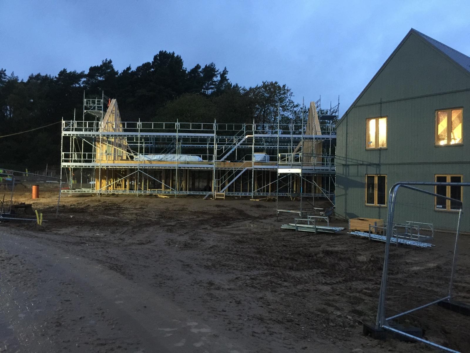 Budowa domu szkieletowego w trakcie realizacji, widoczne rusztowania i konstrukcja drewniana, częściowo obłożona płytami, oświetlone okna w gotowej części budynku, teren budowy z błotem.