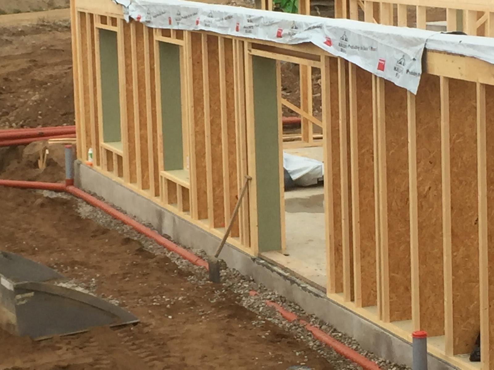Wznoszenie szkieletu drewnianego domu: widoczne pionowe i poziome elementy konstrukcyjne z płyt OSB, izolacja w otworach okiennych, fundament z instalacją zewnętrzną, ziemia wokół budynku.