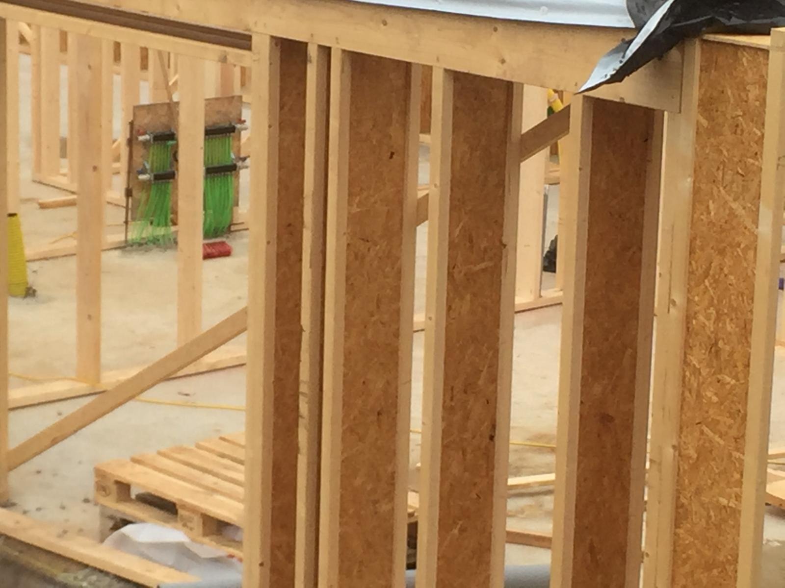 Konstrukcja szkieletowa domu w trakcie budowy, widoczne drewniane belki, płyta OSB, instalacje elektryczne i paleta transportowa w tle.