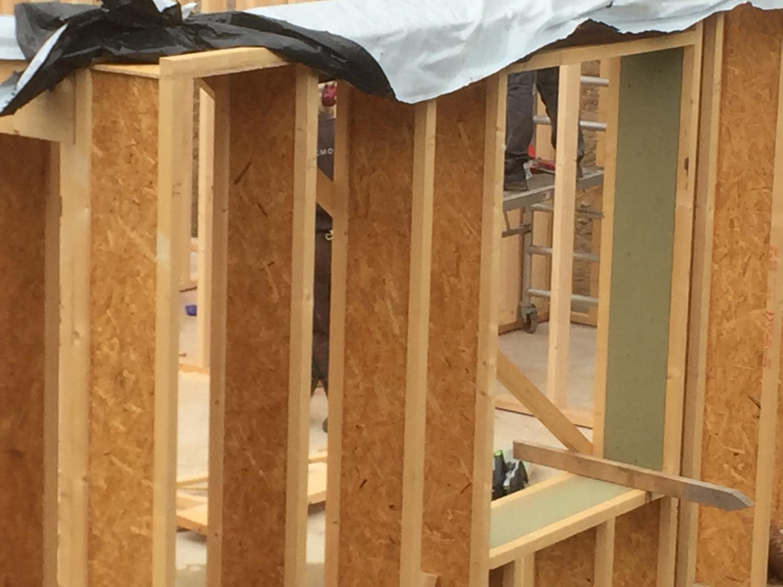 Konstrukcja ścian szkieletowych w trakcie montażu, widoczne drewniane ramy i płyty OSB, fragment izolacji w oknie, w tle drabina i osoba na podwyższeniu.