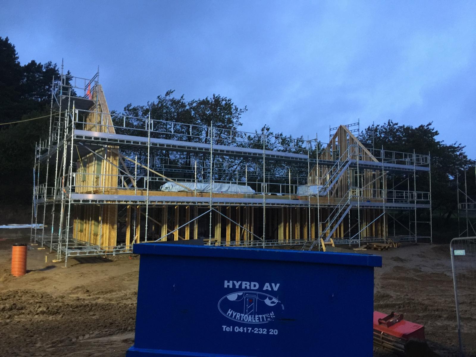 Wznoszenie dwukondygnacyjnego budynku w konstrukcji szkieletowej, otoczonego rusztowaniami, z widocznym niebieskim kontenerem na odpady i pomarańczową rurą kanalizacyjną na placu budowy, przy...