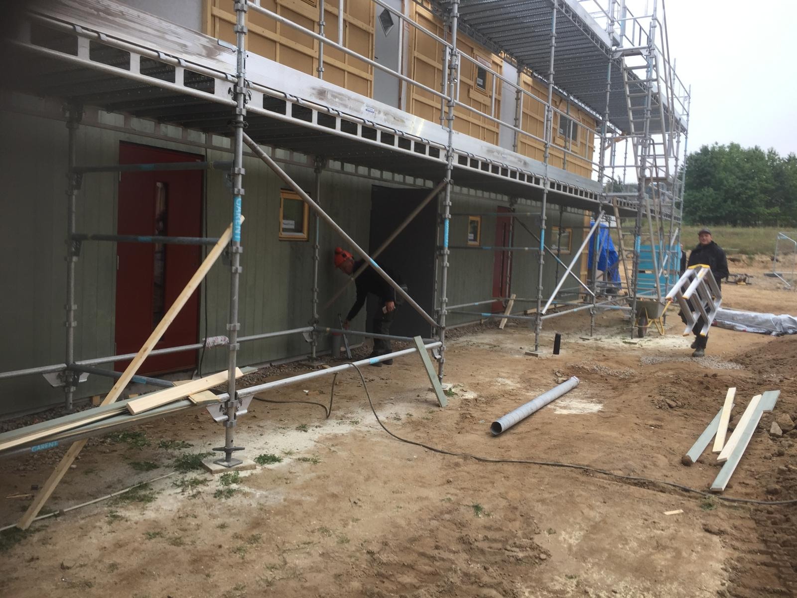 Budowa domu szkieletowego w trakcie: pracownik obrabia ścianę zewnętrzną spod rusztowania, widoczne drewniane elementy konstrukcyjne i narzędzia pracy.