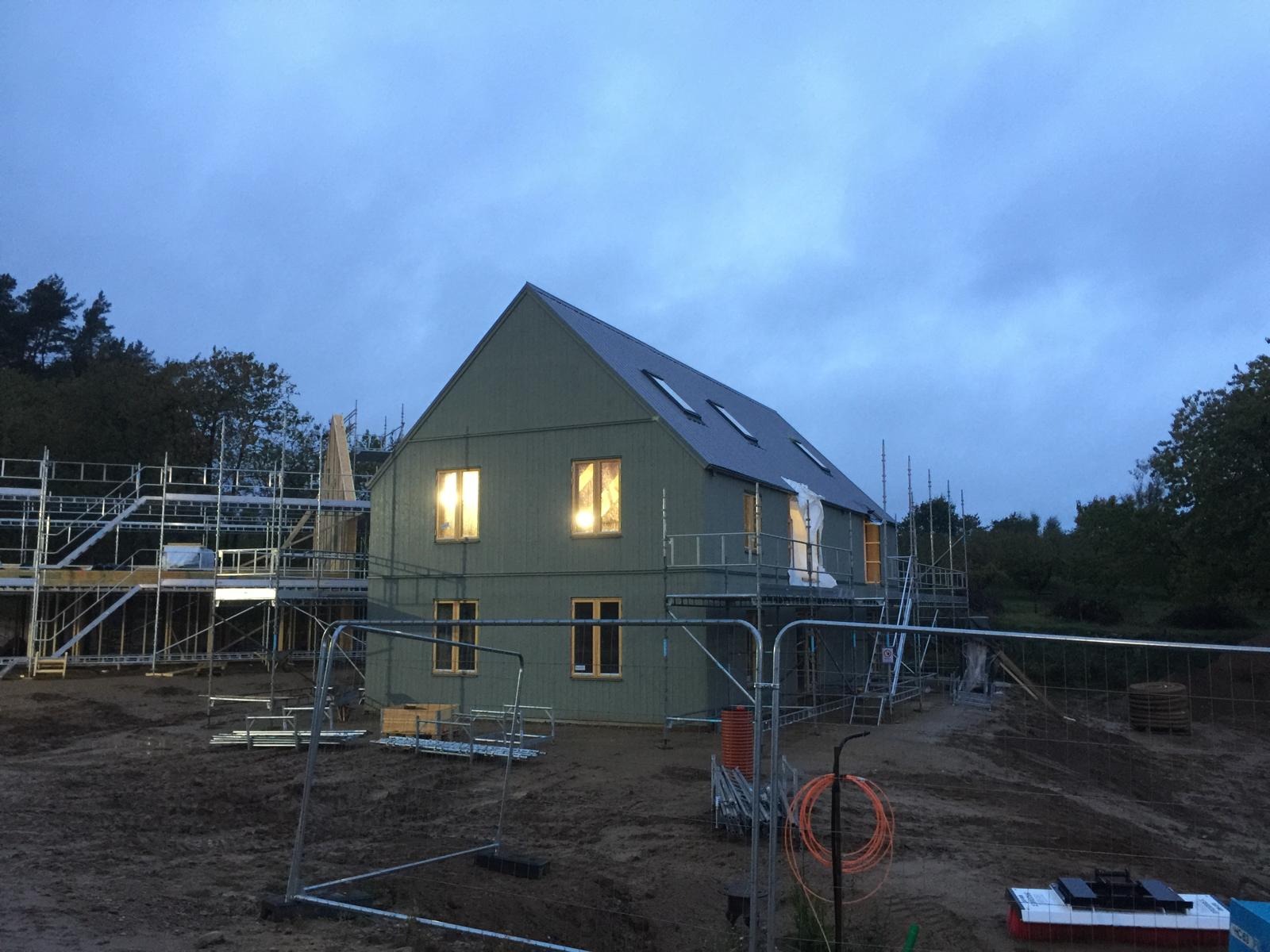 Wczesnym wieczorem szkieletowy dom w trakcie budowy, otoczony rusztowaniami i tymczasowym ogrodzeniem, z oświetlonymi wnętrzami widocznymi przez okna.