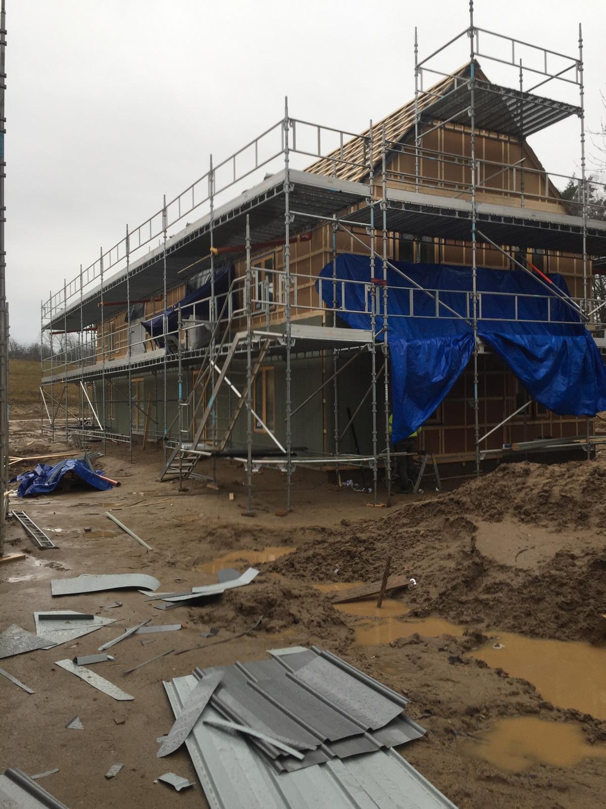 Budowa domu szkieletowego w trakcie: widoczne rusztowania, konstrukcja drewniana, fragmenty niebieskiej plandeki ochronnej i błotniste podłoże z porozrzucanymi elementami blachy.