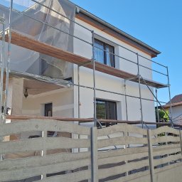 SZYMPRO - Dwupiętrowy dom w trakcie odświeżania elewacji, z rusztowaniem i siatką ochronną, widoczny zza betonowego ogrodzenia.