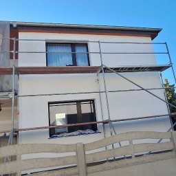 SZYMPRO - Elewacja domu jednorodzinnego w trakcie malowania, widoczne rusztowania i częściowo zakryta siatką ochronną, świeżo pomalowana na biało ściana.