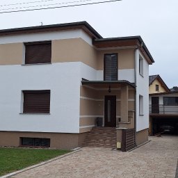 SZYMPRO - Dwukondygnacyjny dom jednorodzinny z jasną elewacją, brązowymi roletami okiennymi i wykończonym wejściem z kolumnami, widoczny z poziomu chodnika z kostki brukowej.