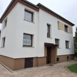 Dwupiętrowy dom z białą elewacją i brązowym cokołem, widoczny z boku, z fragmentem brukowanego podjazdu i zielenią w tle; widoczne pojemniki po farbie i drabina.