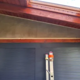 Ściana pod skosem po nałożeniu szarej warstwy gładzi, widoczne drewniane elementy konstrukcyjne dachu i fragment drabiny oparty o roletę zewnętrzną.