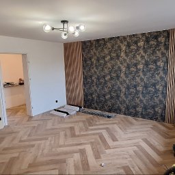 SZYMPRO - Pokój po remoncie: parkiet w jodełkę, ściana z panelami dekoracyjnymi i tapetą w abstrakcyjny wzór, nowoczesna lampa sufitowa, kartony po panelach podłogowych.