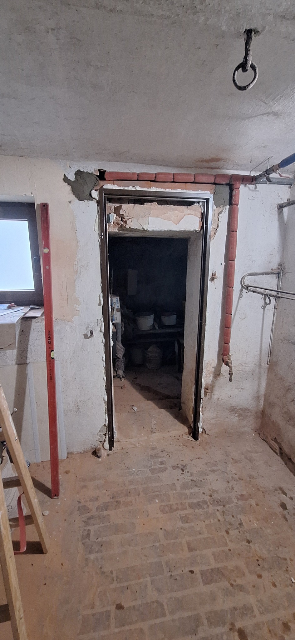 Przygotowanie do montażu ościeżnicy w surowym wnętrzu, widoczne ceglane obramowanie i fragment ściany z resztkami tynku, poziomica oparta o ścianę.