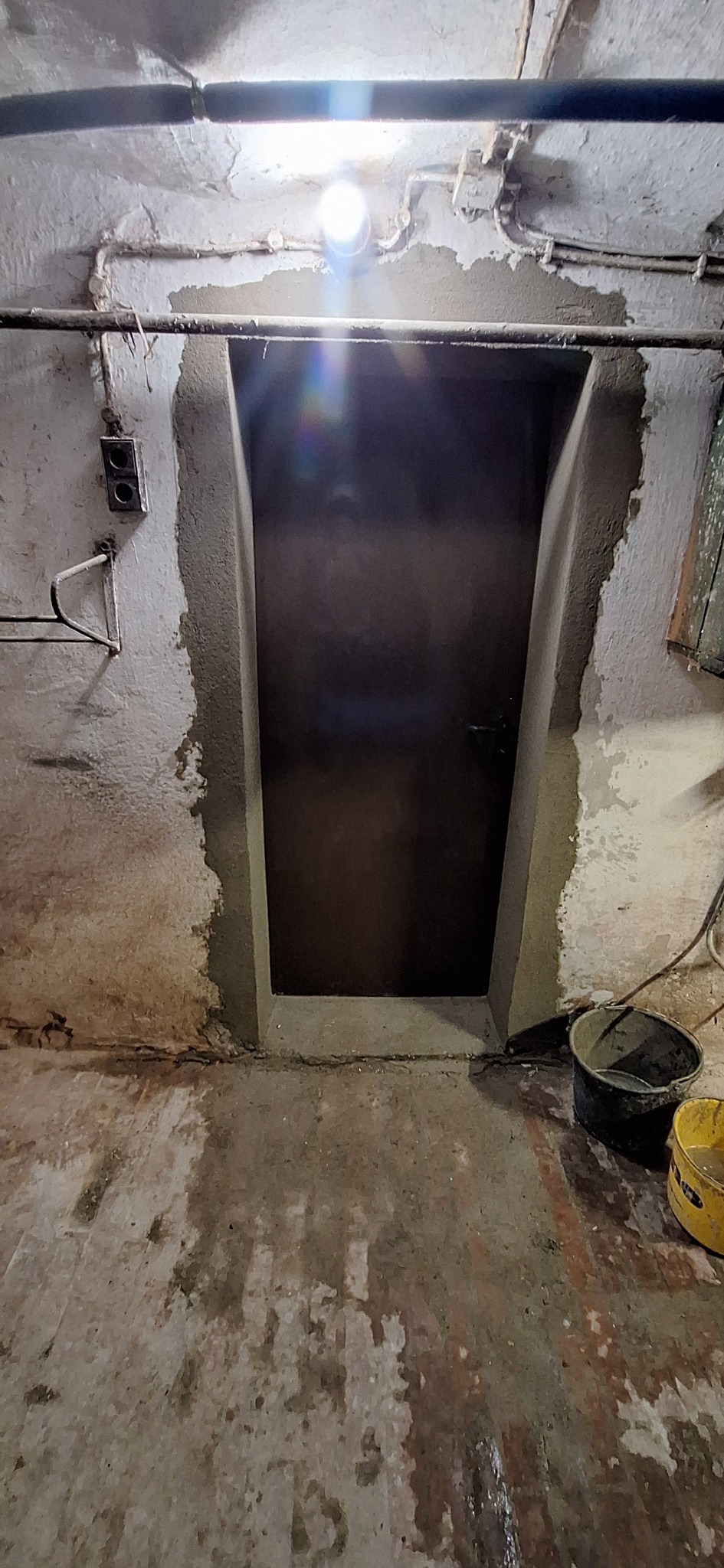 Przygotowanie ościeżnicy drzwiowej do osadzenia z widocznym obrobieniem murarskim, surowym wykończeniem ścian i podłogi oraz instalacjami pod sufitem.