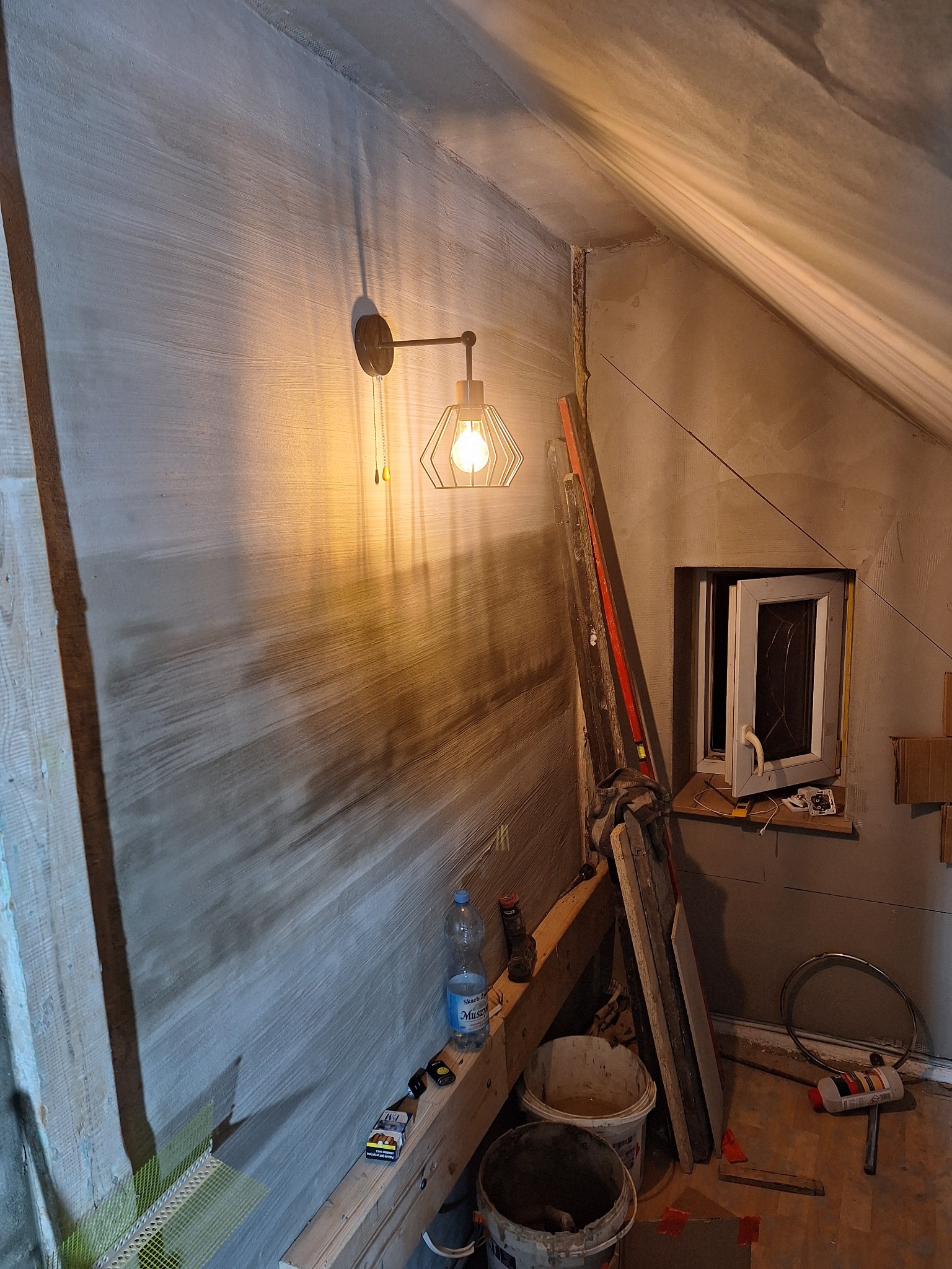 Ściana poddasza w trakcie prac wykończeniowych: świeżo nałożona warstwa szpachli z widocznymi smugami, oświetlona lampą ścienną w stylu industrialnym, z widocznymi narzędziami i materiałami...