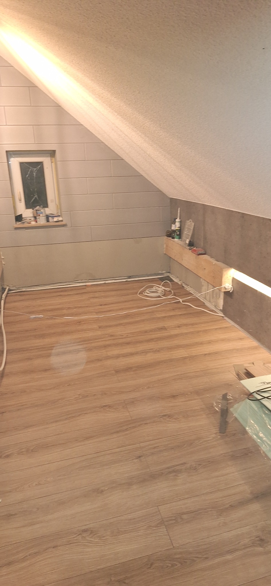 Poddasze po remoncie: panele podłogowe, ściany wyłożone płytkami imitującymi cegłę, małe okno, półka z materiałami budowlanymi i przewody elektryczne na podłodze.