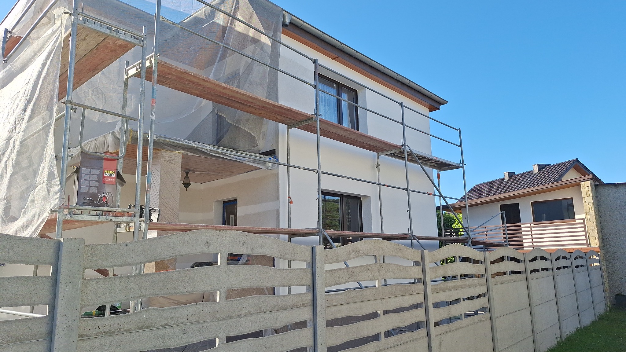 Dwupiętrowy dom w trakcie odświeżania elewacji, z rusztowaniem i siatką ochronną, widoczny zza betonowego ogrodzenia.