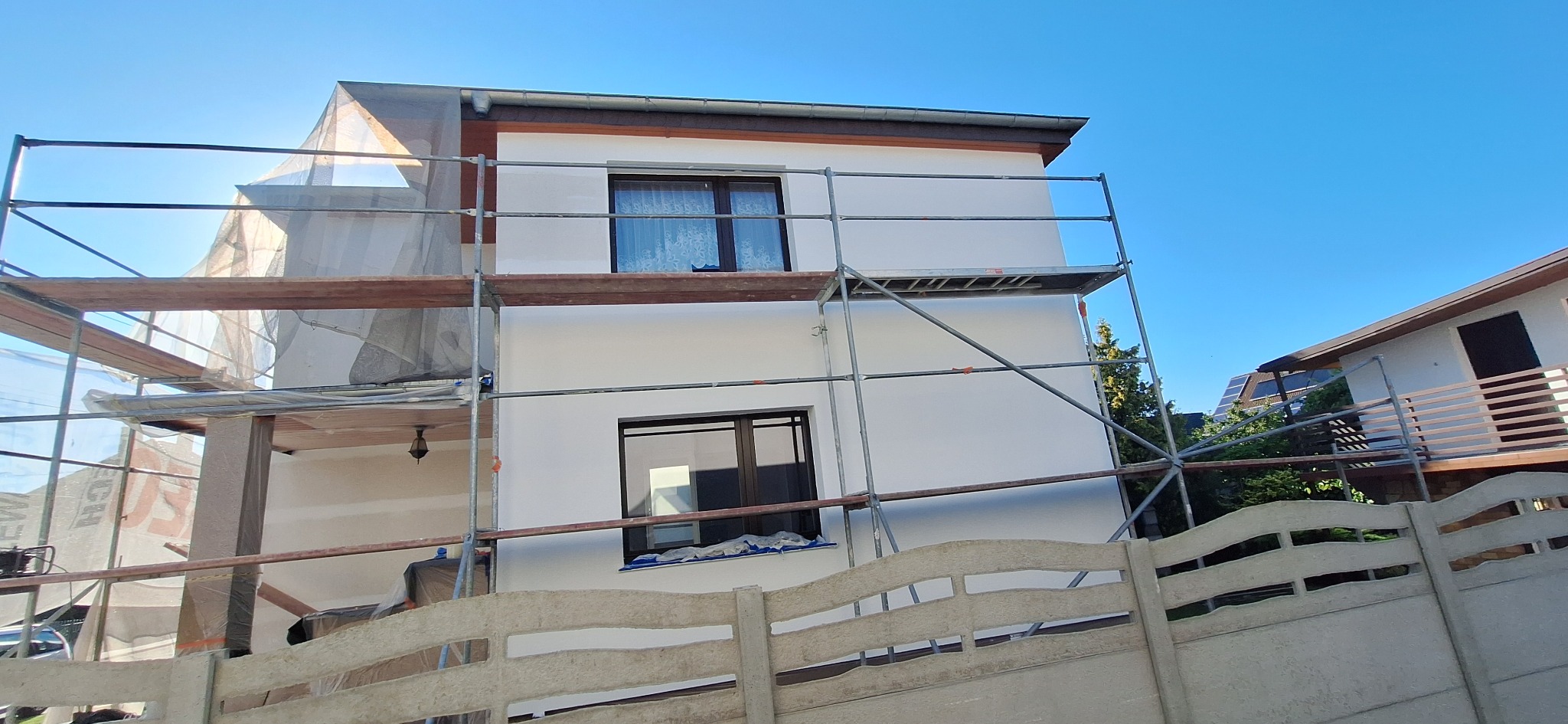 Elewacja domu jednorodzinnego w trakcie malowania, widoczne rusztowania i częściowo zakryta siatką ochronną, świeżo pomalowana na biało ściana.