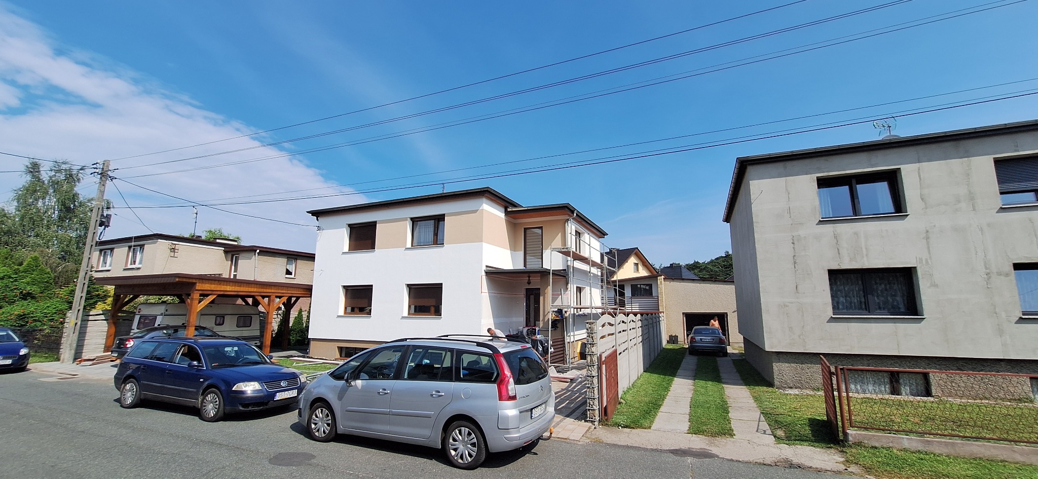 Dwukondygnacyjny dom jednorodzinny w trakcie malowania elewacji, widoczne rusztowania, z boku zaparkowane dwa samochody osobowe.