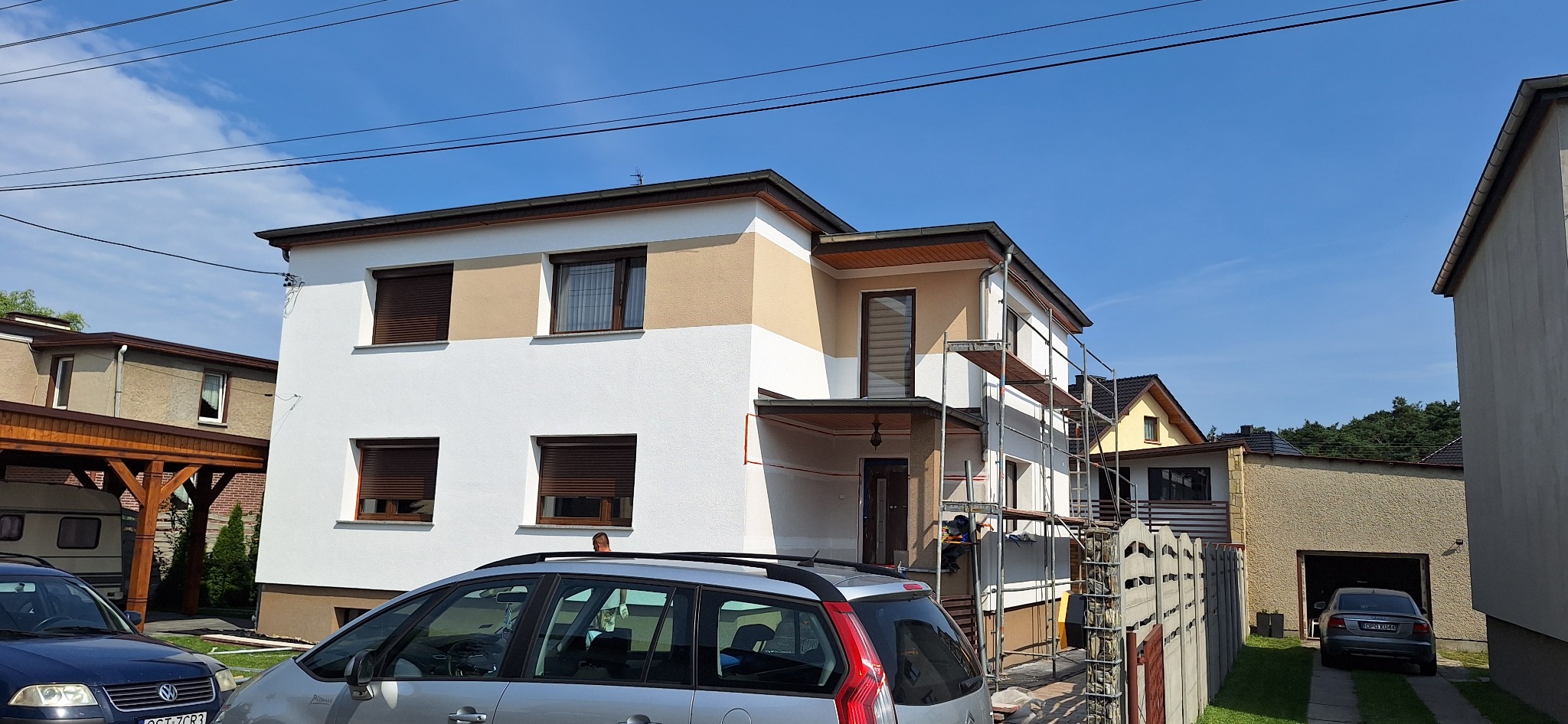 Dwukondygnacyjny dom jednorodzinny w trakcie prac elewacyjnych, z widocznymi rusztowaniami i fragmentem samochodu osobowego na pierwszym planie.