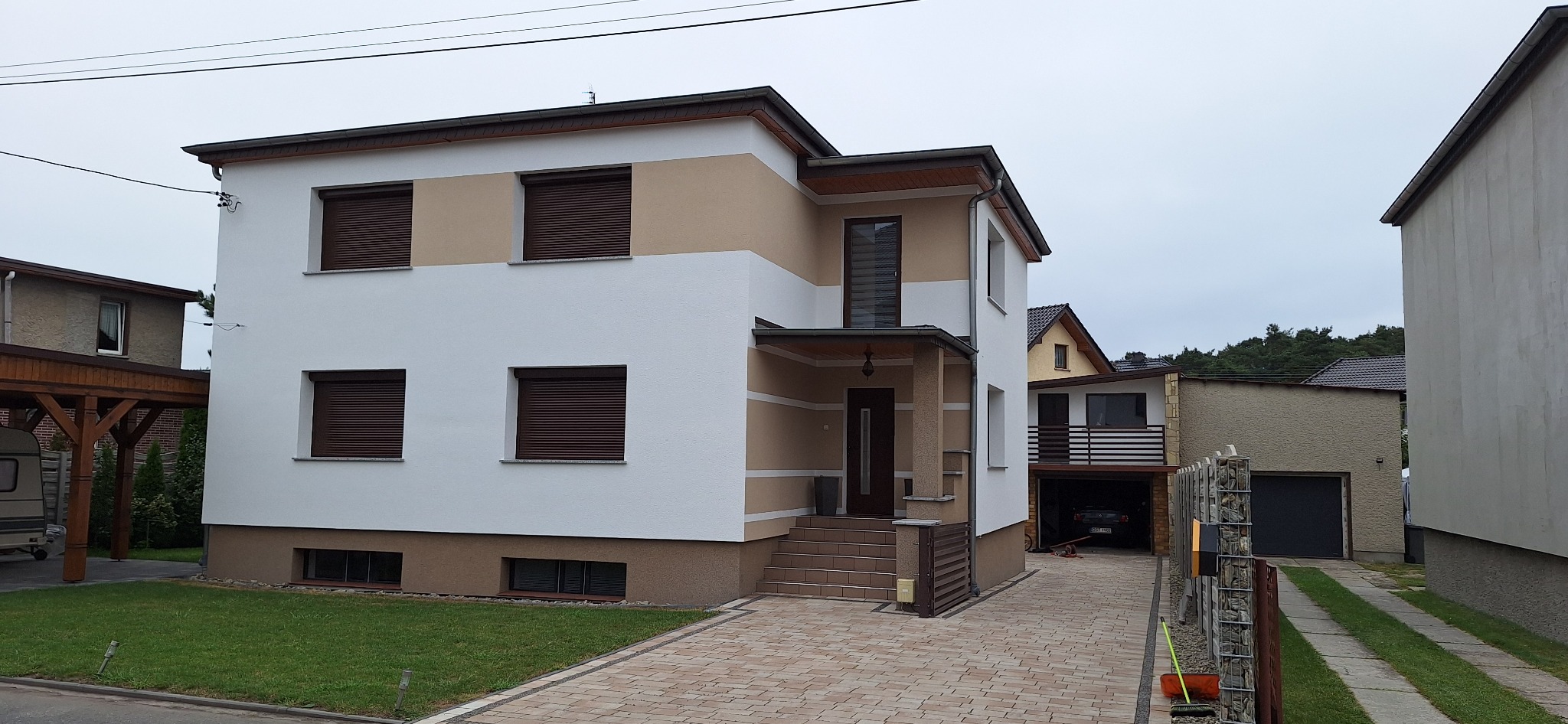 Dwukondygnacyjny dom jednorodzinny z jasną elewacją, brązowymi roletami okiennymi i wykończonym wejściem z kolumnami, widoczny z poziomu chodnika z kostki brukowej.