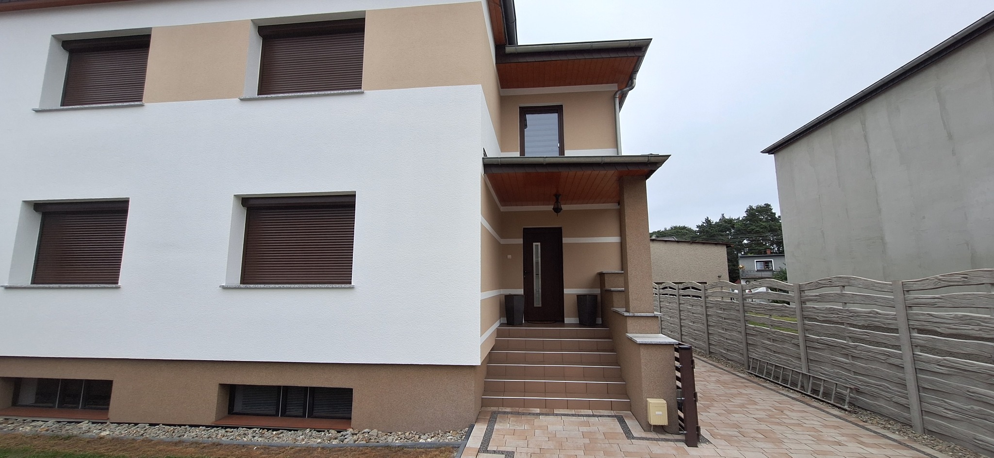Elewacja dwukondygnacyjnego domu z brązowymi roletami zewnętrznymi, beżowo-biała fasada, wejście z brązowymi drzwiami i schodami wyłożonymi płytkami, widok z poziomu chodnika.