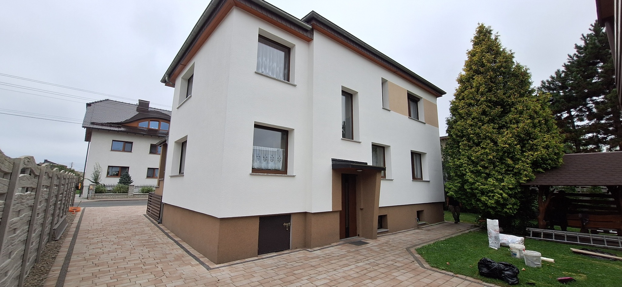 Dwupiętrowy dom z białą elewacją i brązowym cokołem, widoczny z boku, z fragmentem brukowanego podjazdu i zielenią w tle; widoczne pojemniki po farbie i drabina.