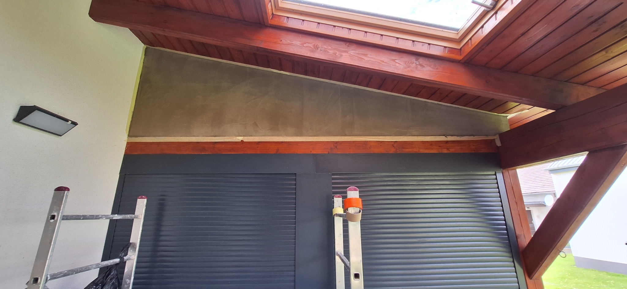 Ściana pod skosem po nałożeniu szarej warstwy gładzi, widoczne drewniane elementy konstrukcyjne dachu i fragment drabiny oparty o roletę zewnętrzną.