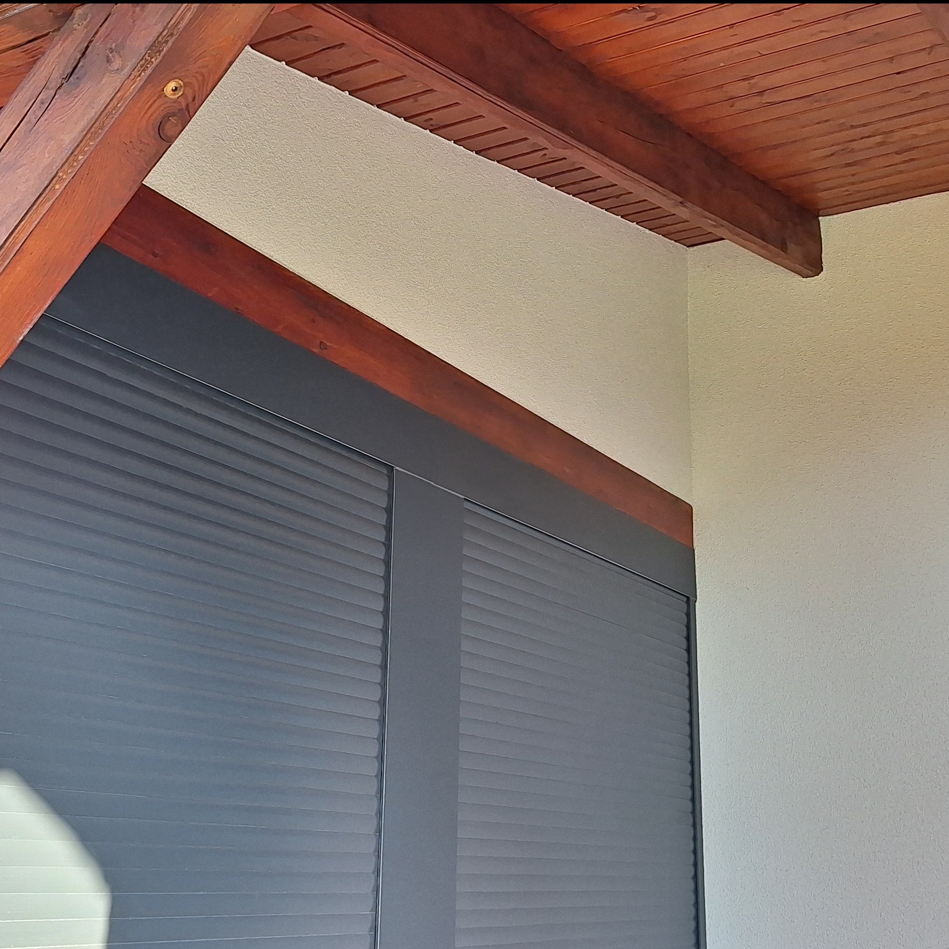 Szare rolety zewnętrzne zamontowane pod drewnianą konstrukcją dachu na tle jasnej ściany o ziarnistej fakturze.