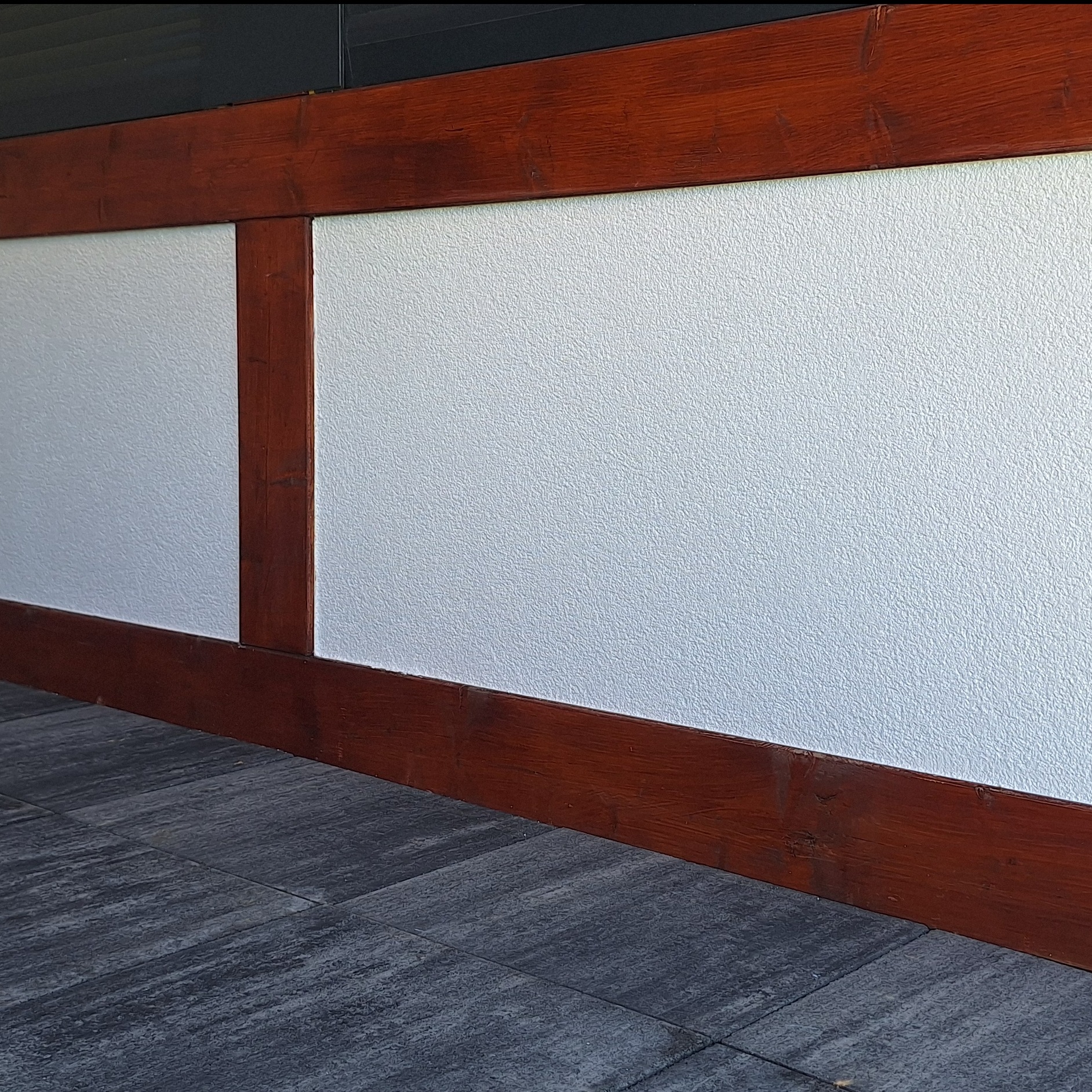 Wykończona ściana z dekoracyjnym tynkiem w kolorze białym, obramowana ciemnobrązową drewnianą konstrukcją i szarą kamienną posadzką.