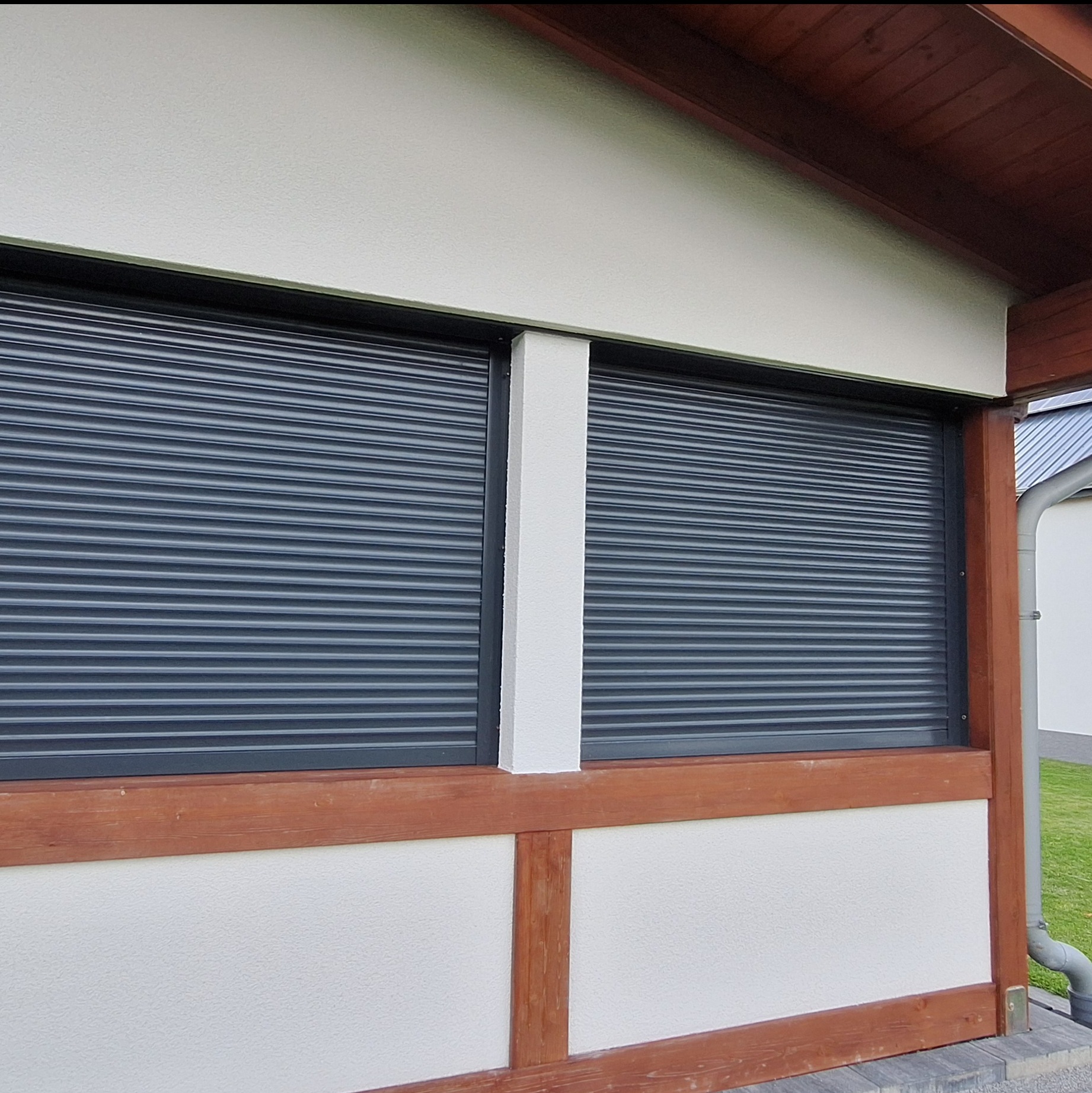 Zamknięte, szare rolety zewnętrzne w oknach budynku z białą elewacją i drewnianą konstrukcją nośną.