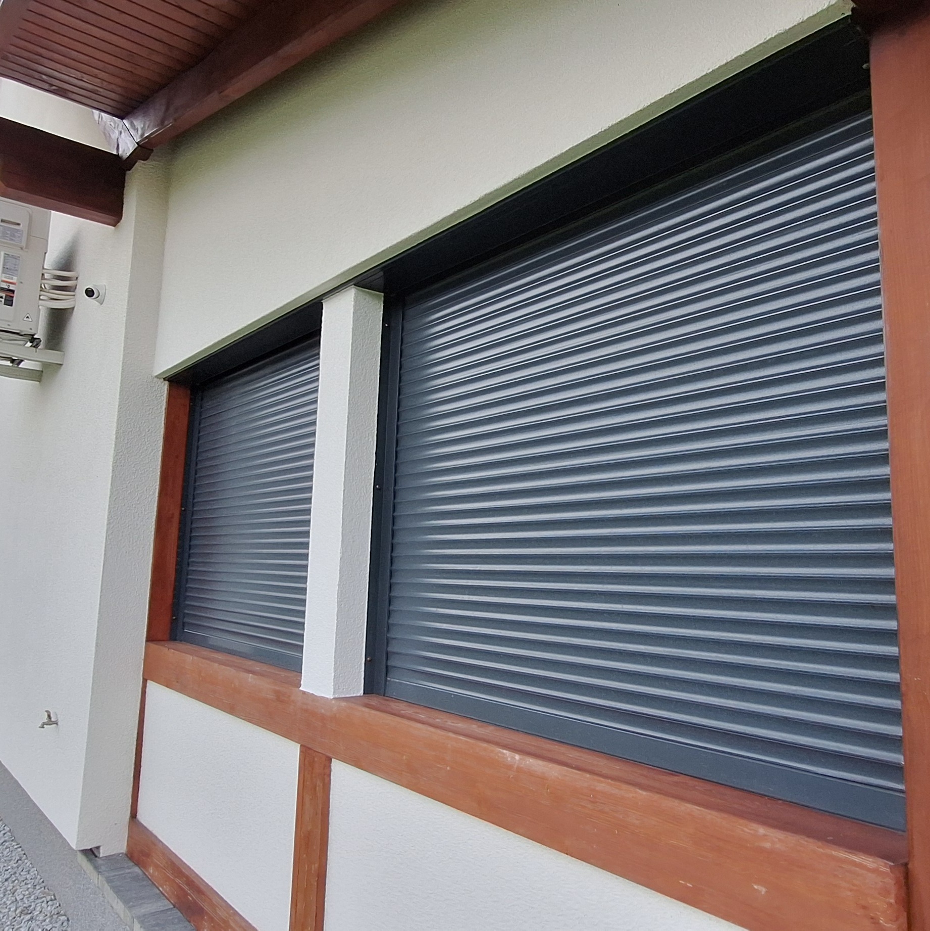 Dwa okna z opuszczonymi, szarymi roletami zewnętrznymi, widoczne z zewnątrz budynku z białymi ścianami i drewnianą ramą okienną w kolorze brązowym.