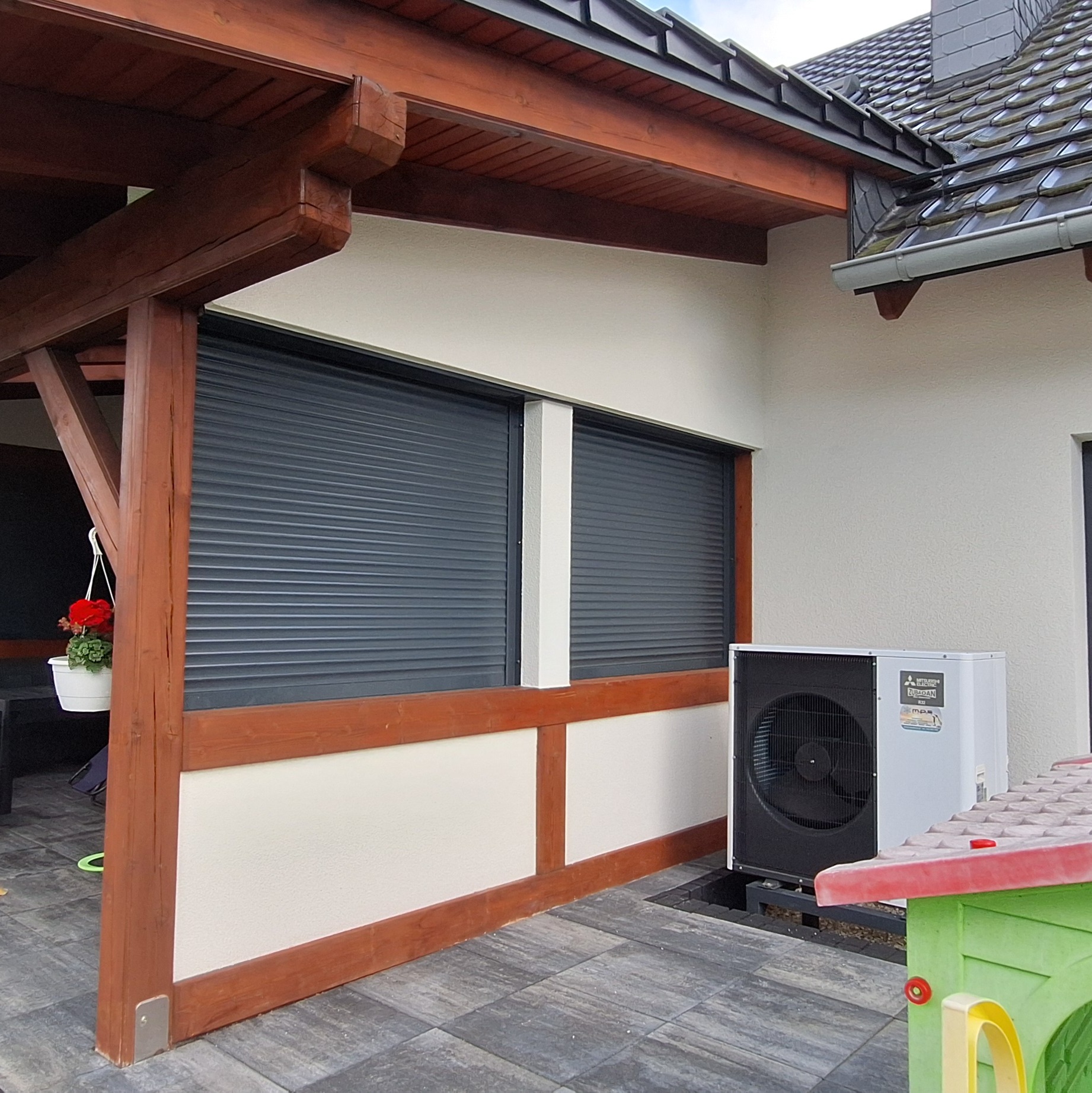 Biała ściana budynku z dwoma oknami zabezpieczonymi roletami zewnętrznymi w kolorze grafitowym, drewniana konstrukcja z belek w kolorze brązowym, zewnętrzna jednostka klimatyzacji Mitsubishi...