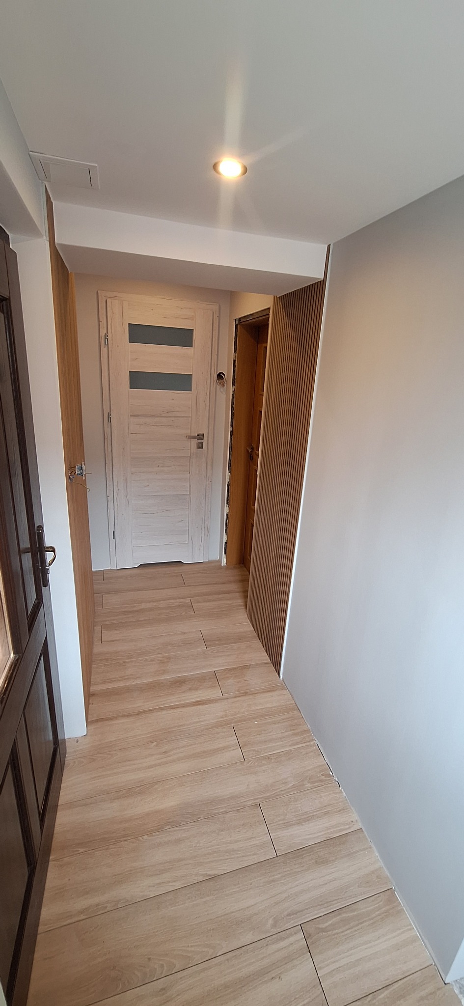 Wnętrze korytarza po remoncie: jasne płytki podłogowe imitujące drewno, białe ściany i zamontowane drzwi wewnętrzne z drewnianymi lamelami.