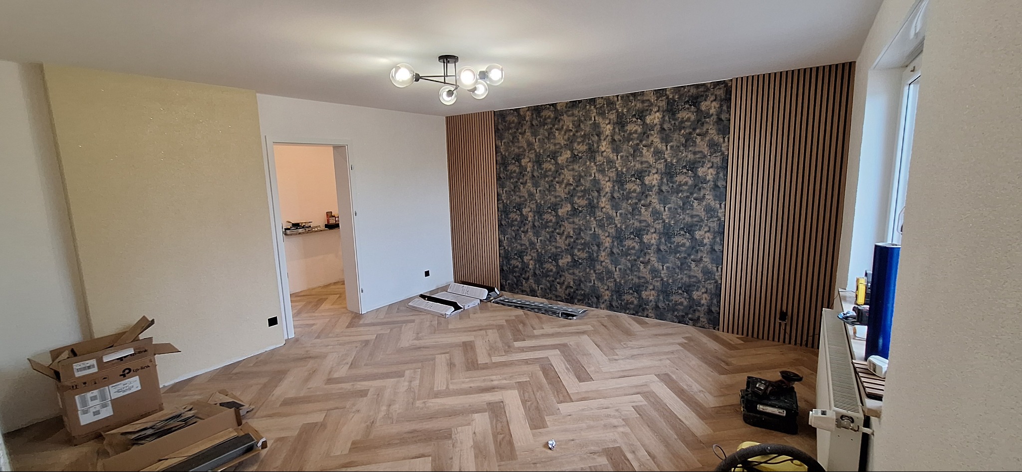 Pokój po remoncie: parkiet w jodełkę, ściana z panelami dekoracyjnymi i tapetą w abstrakcyjny wzór, nowoczesna lampa sufitowa, kartony po panelach podłogowych.