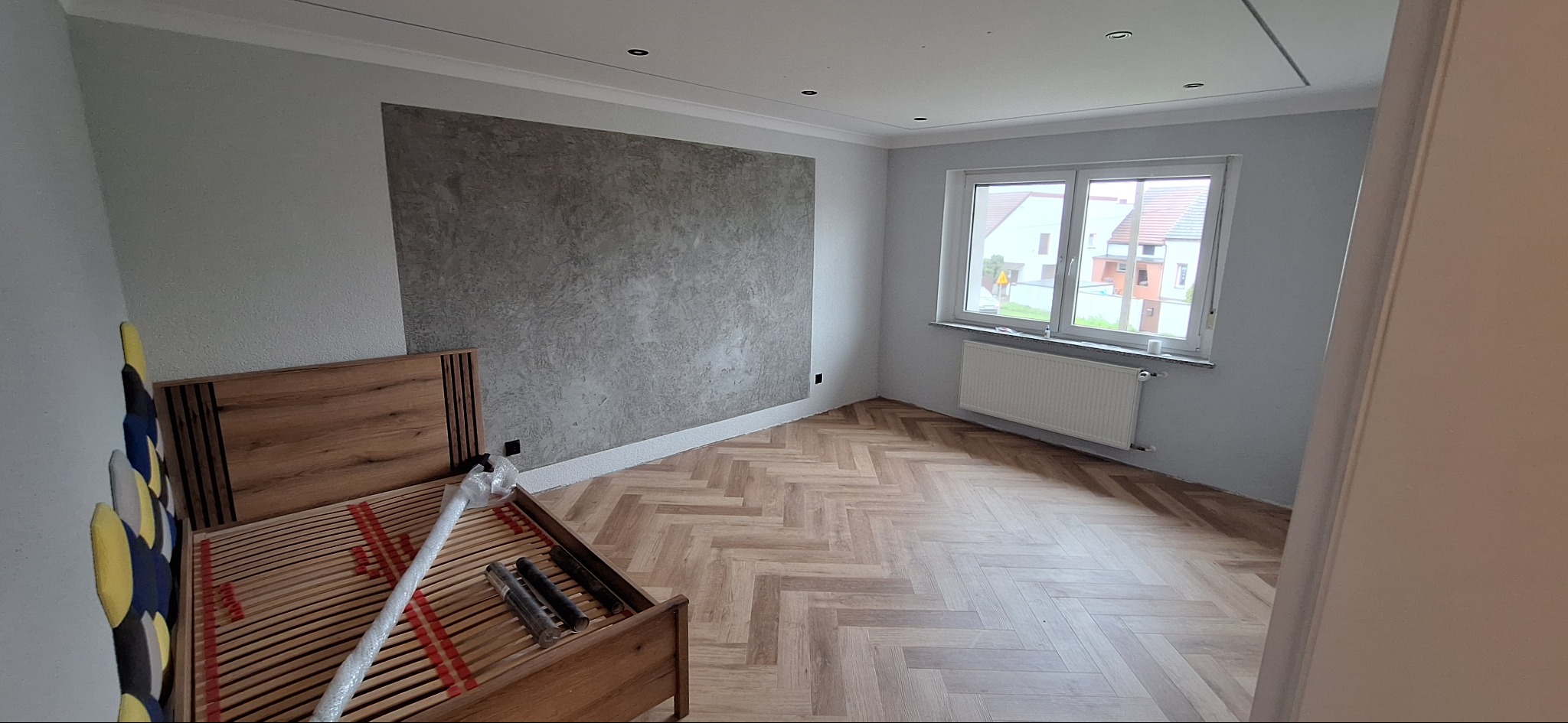 Pokój po remoncie: gładkie, szare ściany, podłoga w jodełkę, okno z grzejnikiem, częściowo widoczne łóżko z drewnianym wezgłowiem. Nowoczesne wykończenie wnętrz.