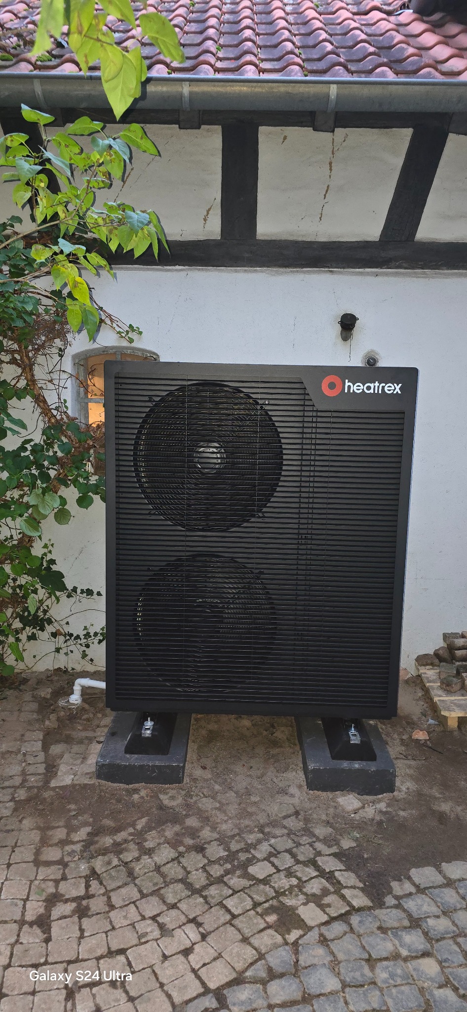 Czarna pompa ciepła Heatrex na betonowych podstawach, zainstalowana przy budynku z murem pruskim. Widoczne detale elewacji i fragment roślinności.