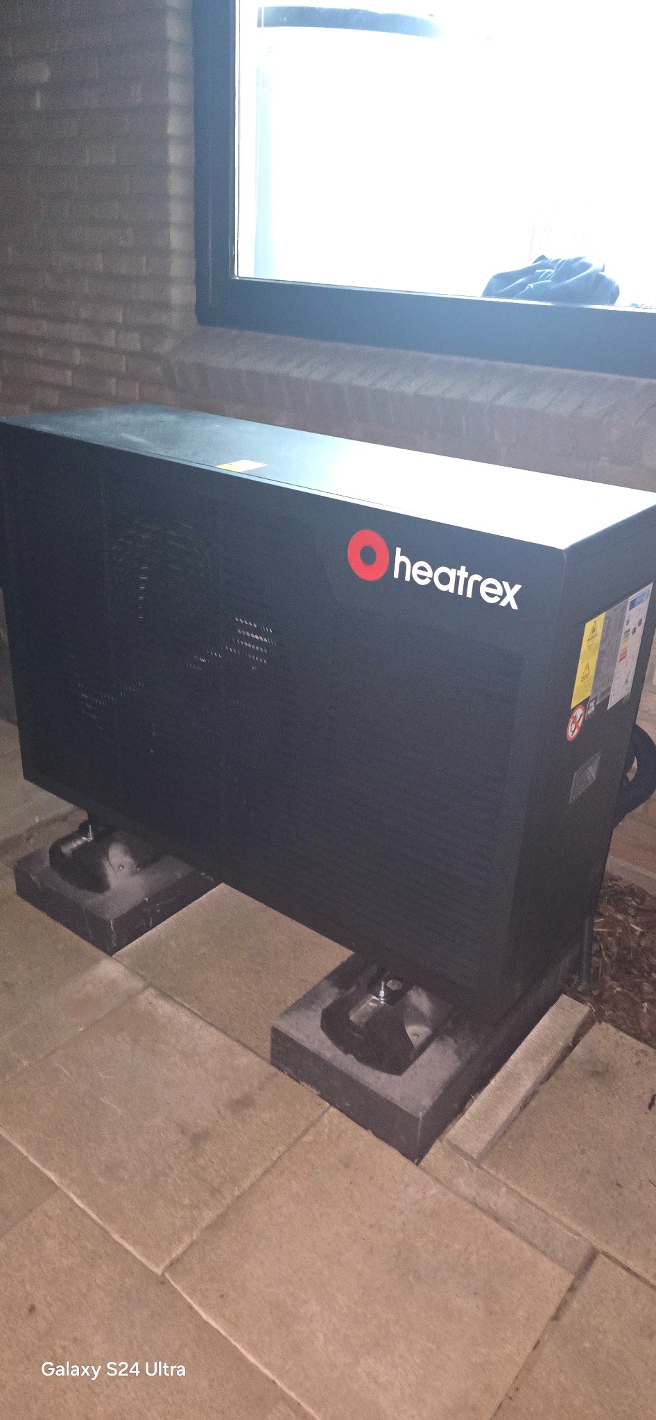 Czarna pompa ciepła Heatrex na betonowych podstawach, ustawiona na zewnątrz budynku, z widocznym oknem w tle.