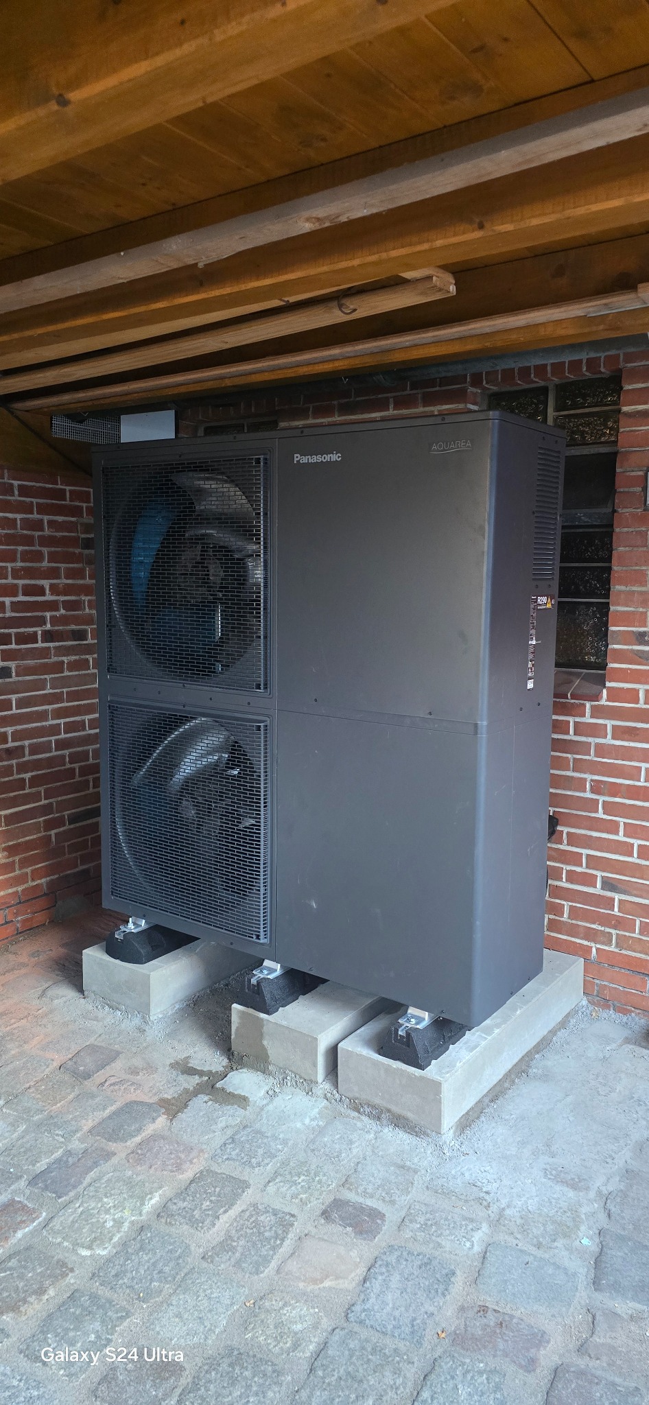 Szara pompa ciepła Panasonic Aquarea, zamontowana na betonowych wspornikach na brukowanym podłożu, przy ceglanej ścianie i drewnianym suficie.