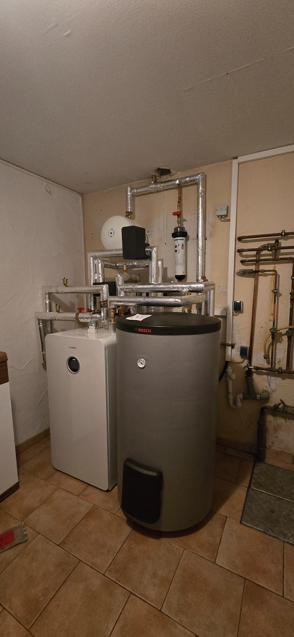 Kompletna instalacja grzewcza Bosch z zbiornikiem i rurami w izolacji, widoczne połączenia i manometry. Całość w pomieszczeniu z płytkami na podłodze.