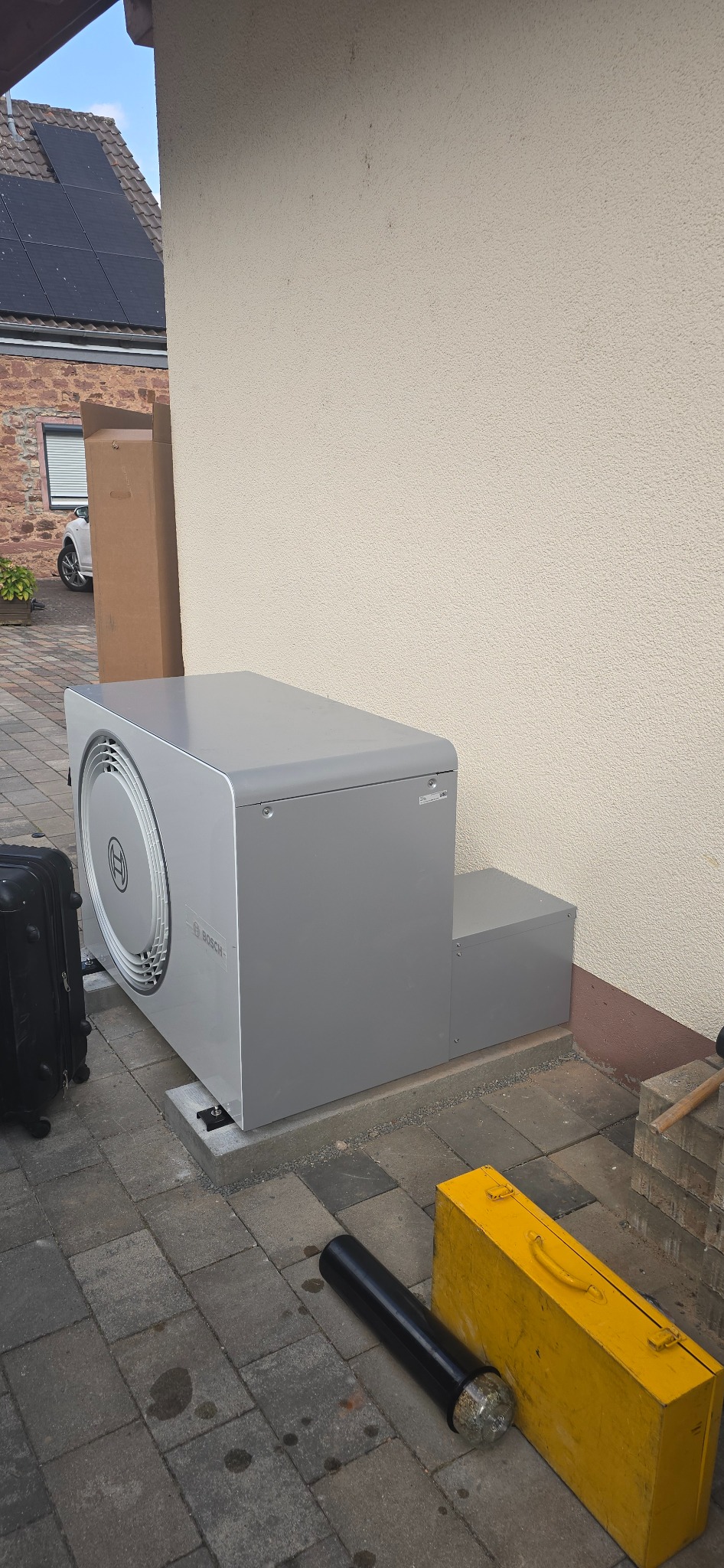 Szara pompa ciepła Bosch zamontowana na zewnątrz budynku, widoczna na tle beżowej ściany i kostki brukowej. Obok leżą narzędzia i walizka.