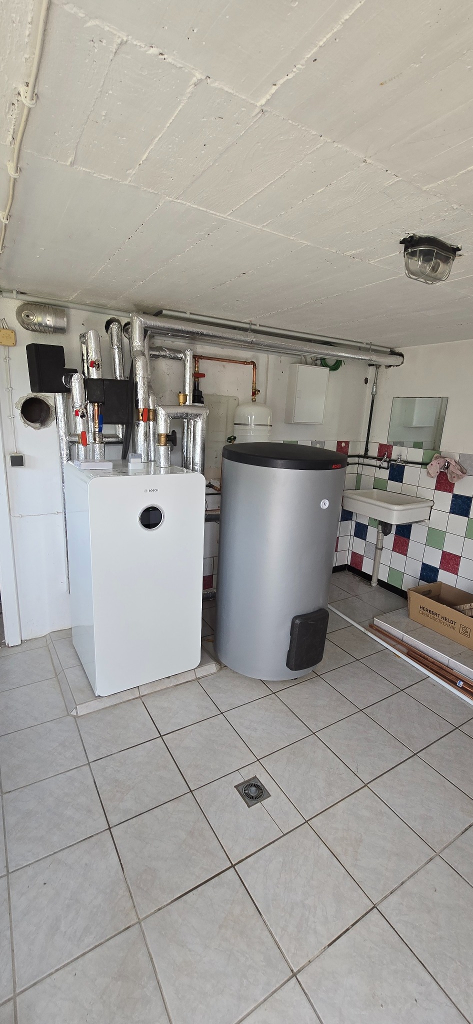 Biała pompa ciepła Bosch z szarym zasobnikiem w pomieszczeniu z białą kratką na podłodze i instalacją grzewczą na ścianie.