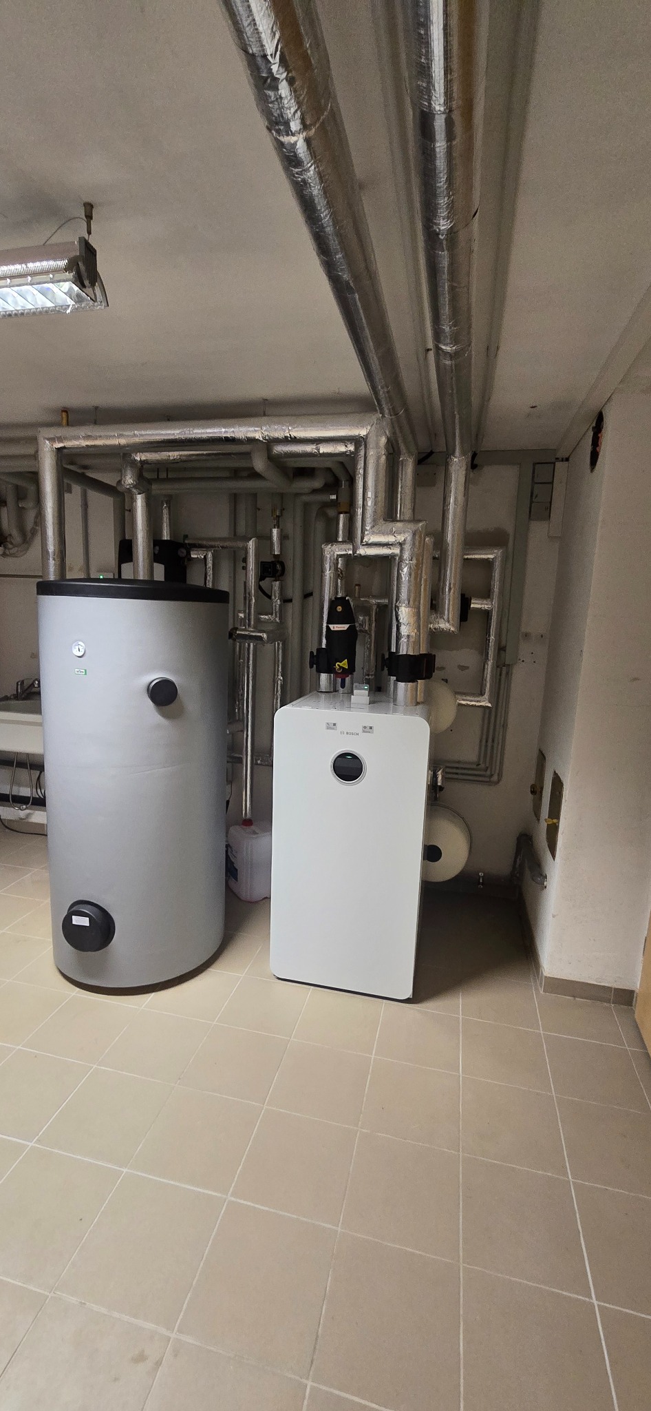 Szara pompa ciepła Bosch obok zbiornika buforowego w kotłowni z rurami na suficie. Widok na instalację w pomieszczeniu technicznym.