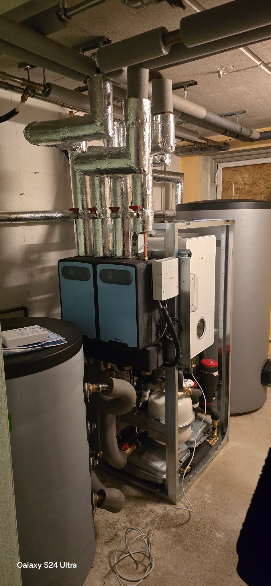 Złożona instalacja grzewcza z niebieskimi panelami Meibes, białym modułem Bosch i srebrnymi rurami izolowanymi folią, w pomieszczeniu technicznym.