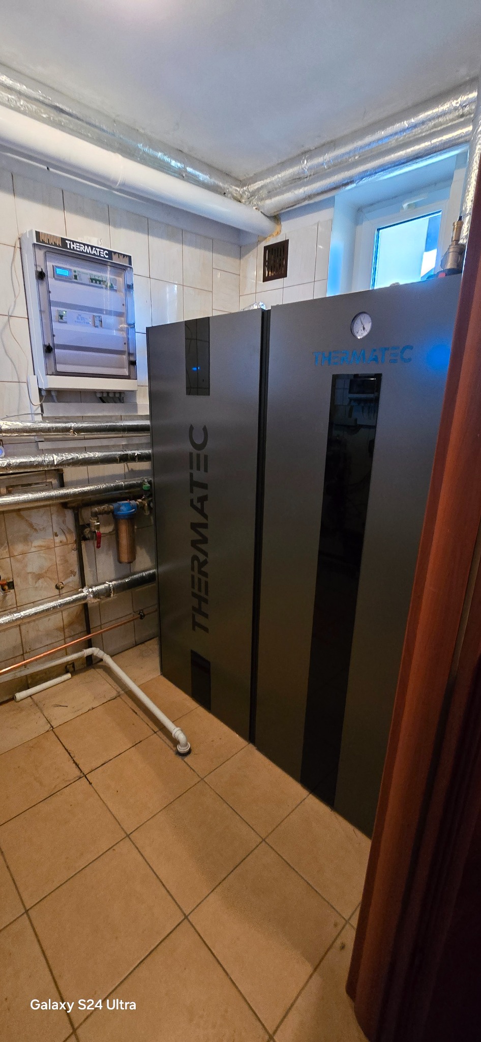 Zainstalowana pompa ciepła Thermatec w kotłowni z białą glazurą. Widoczne rury instalacyjne i panel sterowania na ścianie.