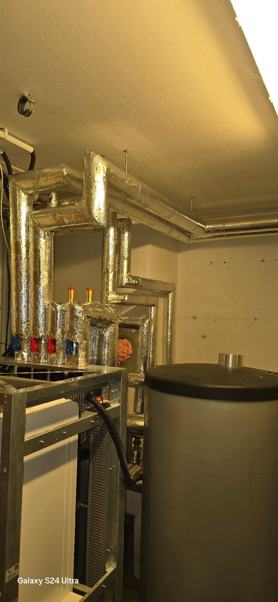 Złożona instalacja grzewcza z rurami w izolacji aluminiowej, zbiornikiem i metalową konstrukcją. Widoczne zawory z oznaczeniami kolorystycznymi.