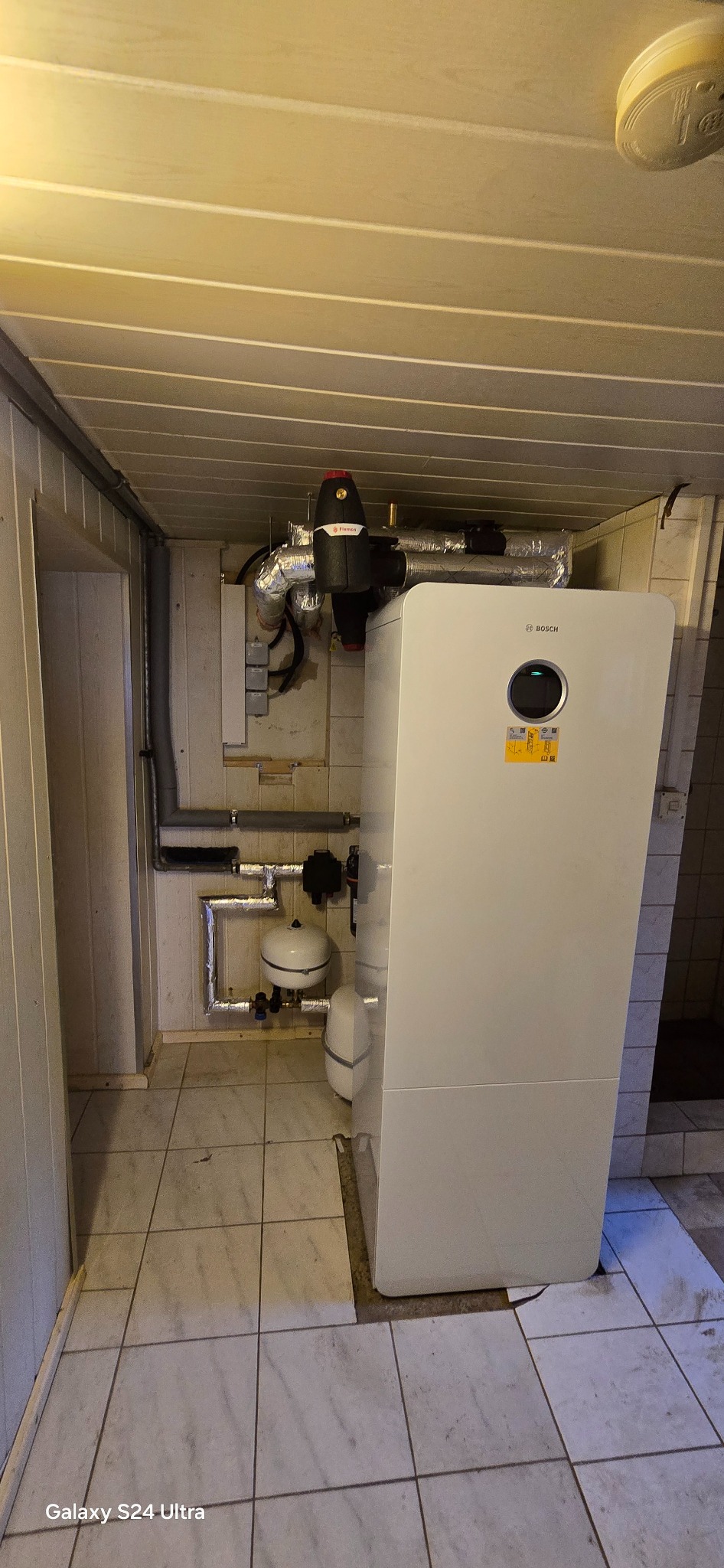 Biała pompa ciepła Bosch w kotłowni w Bielsko-Białej. Widoczne rury, zbiorniki wyrównawcze i czujnik dymu na suficie. Instalacja w pomieszczeniu z płytkami.